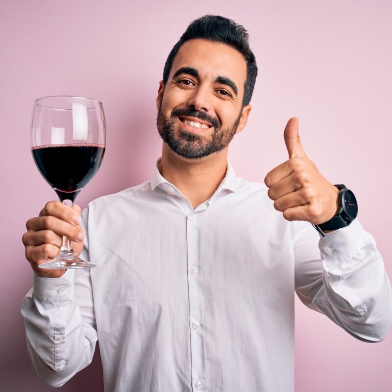 Young handsome man with beard drinking glass of red wine over isolated pink background happy with big smile doing ok sign, thumb up with fingers, excellent sign Young handsome man with beard drinking glass of red wine over isolated pink background happy with big smile doing ok sign, thumb up with fingers, excellent sign