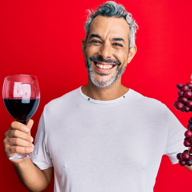 Middle age grey-haired man holding branch of fresh grapes and red wine smiling with a happy and cool smile on face. showing teeth.